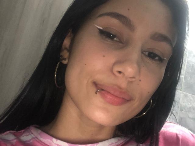 KataleyaMansson has dark brown hair, black eyeliner, a septum piercing, and gold hoop earrings. She's wearing a pink shirt with white patterns, smiling slightly.