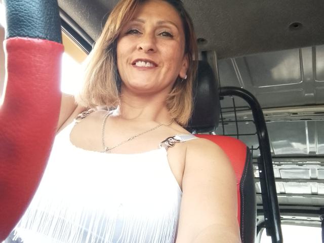CamilaAbbey, smiling with medium-length blonde hair, wearing a white, pleated tank top with silver straps, holding a red armrest, inside a van.