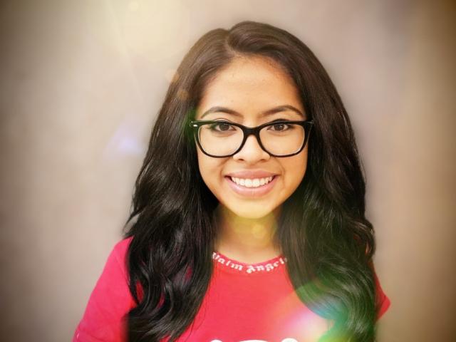 SoniaLegacy with long black hair, wearing black glasses, and a red shirt, smiling at the camera, showing her white teeth, and a subtle lens flare.