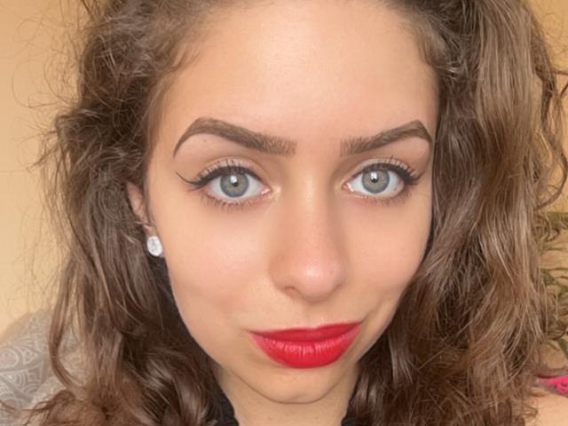 AsterCrystal has blue eyes, red lipstick, and a cute face with curly brown hair. She's wearing diamond earrings and has thick, arched eyebrows. Her ass is not visible.