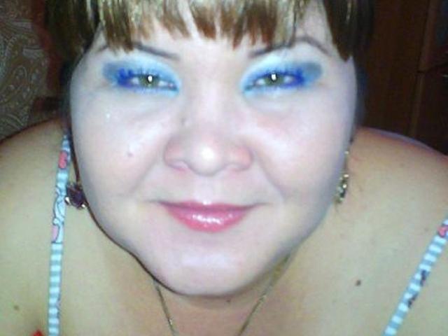 ElizabethFunny with blue eye makeup, short brown hair, pink lips, and a slight smile, showing her chubby face and bare shoulders with a beaded strap.