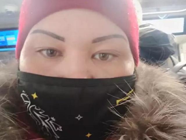 ElizabethFunny has thick eyebrows, brown eyes, and wears a red beanie with a black mask showing her nose and mouth. She's in a furry coat.