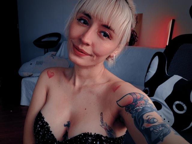 SaraleePoison with blonde bangs, smiling, showing small red heart tattoos on shoulders, black lace bra, and colorful tattoo on right arm, in dimly lit room.