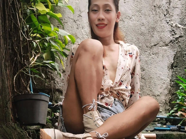 Darcy, zomerse outfit met de knieën omhoog, draagt een bloemenprint blouse met strik, denim shorts en veterschoenen, lachend, met een subtiele glimp van haar borsten, zittend op een groene, begroeide plek buiten..