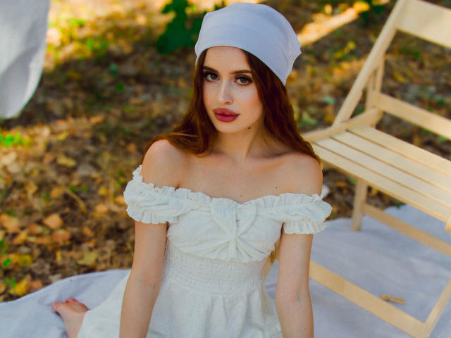 CherylFoxy with long brown hair, white off-shoulder dress, and white headscarf, sitting on white cloth, wooden chair in background, looking up with red lipstick, bare shoulders.