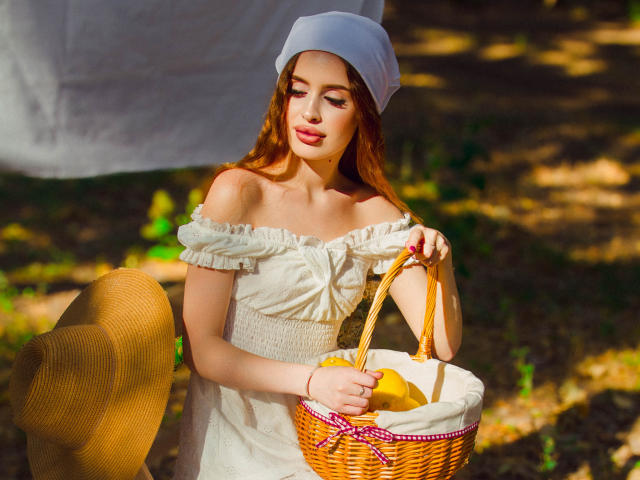 CherylFoxy with red hair, white off-shoulder dress, and white headscarf, holding wicker basket with yellow apples, standing beside yellow hat, sunny forest background.
