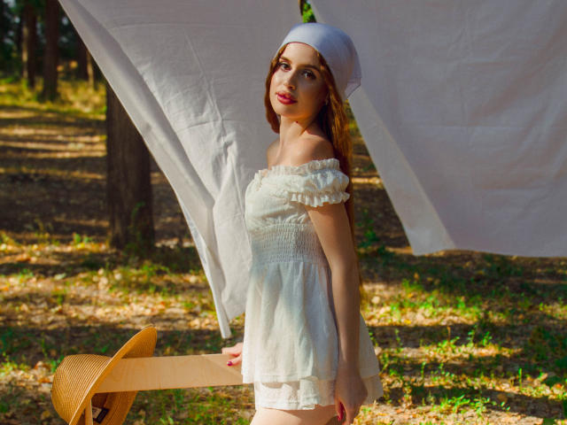 CherylFoxy with light brown hair, white off-shoulder dress, and white headscarf, holding a straw hat, standing under a white sheet, in a sunny forest. Boobs visible.