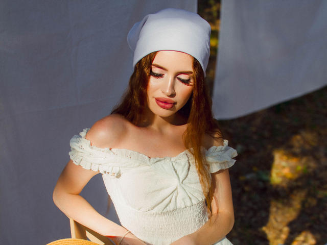 CherylFoxy with long brown hair, white off-shoulder dress, and white cap, sitting, looking down, sunlit, small tits, cute face, red lipstick, soft light, daytime, outdoors.