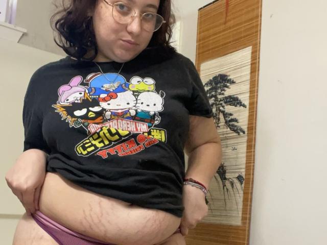 PinkBerrieBabe with glasses, wearing a black "Hello Kitty" t-shirt, lifting it to show her chubby belly and stretch marks, wearing pink panties, in a room.
