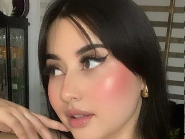 LaskmyEvans has pink blushed cheeks, dark eyeliner, and long black eyelashes, with her hand near her face, wearing gold earrings, and looking to the side.