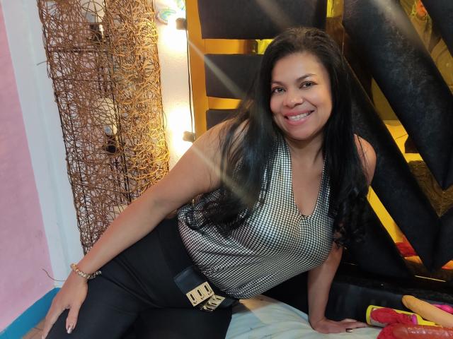 HellenaSexy, sorrindo com longos cabelos pretos, vestindo uma blusa sem mangas xadrez e calças pretas, apoiada em uma cama com uma parede amarela e preta atrás dela..