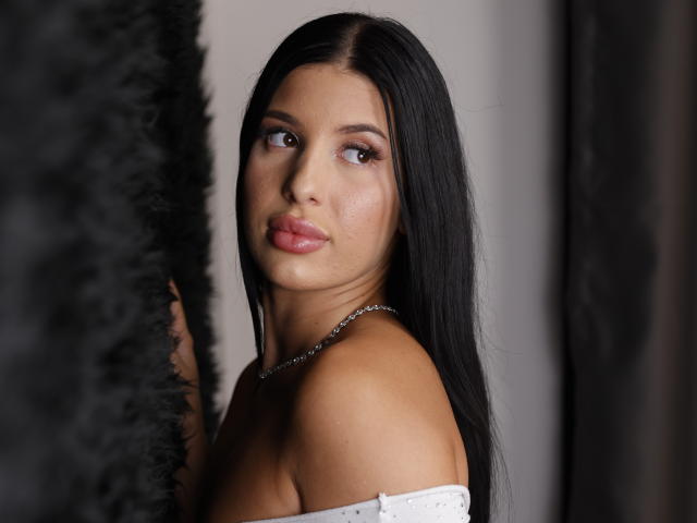 TiffanyRides with long black hair, looking to the side, has pink lips and a shiny nose, wearing an off-shoulder white top, necklace, and no visible boobs or ass.