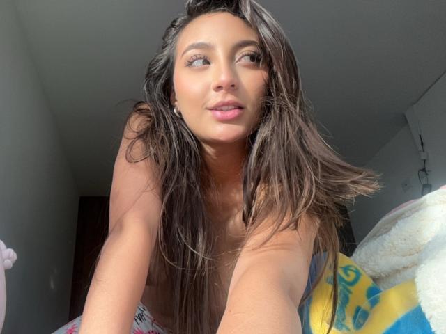 DianaFernandezz with long brown hair, looking up with a slight smile, pink lips, and tan skin, topless, sunlight on her face, arms forward, blue and yellow blanket.