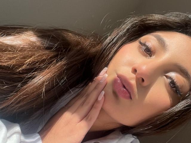 DianaFernandezz has long brown hair, full pink lips, and is gazing slightly upwards with a hand near her face. She has a sexy, soft look with sunlit skin.