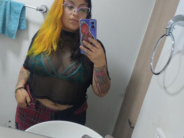 GineBralkino con cabello amarillo y negro, gafas, top negro transparente, sujetador de encaje verde, pantalones a cuadros, tatuajes, sosteniendo un teléfono morado, selfie frente al espejo, baño blanco, toalla azul, anillo..