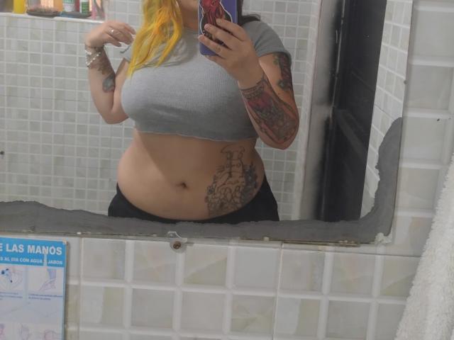 GineBralkino met blond haar, een grijze top, zwarte broek, een getatoeëerde arm, die een telefoon vasthoudt, een spiegelzelfportret, waarbij haar buik en grote borsten zichtbaar zijn, een witte betegelde badkamer, een zwarte spiegelrand..