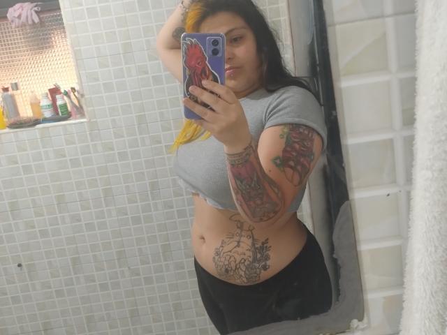 GineBralkino with tattoos, holding phone, wearing grey crop top, black pants, in bathroom mirror selfie, showing belly tattoo, arm tattoos, and medium tits. Long black hair, yellow streak.