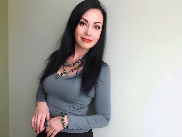 RoxolanaXDrim has black hair, a grey long-sleeve top, red lipstick, and multiple necklaces with beads. She's smiling, with hands resting on her black skirt.