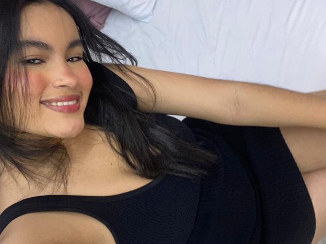 MiiaPalmer con una canottiera nera, sorride alla telecamera, sdraiata sul letto, mostrando il seno, capelli neri, fard rosa, cuscino bianco sullo sfondo, braccio destro appoggiato..