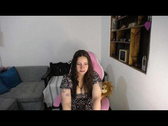 LorelayIncanto with medium tits, brown wavy hair, zebra print dress, sitting in pink chair, arms resting on desk, slight smile, gray sofa, stuffed lion in background.