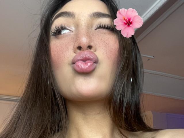 DianaFernandezz with puffy pink lips, winking, dark brown hair, a pink hibiscus flower in her hair, and freckled skin, wearing hoop earrings, looking cute.