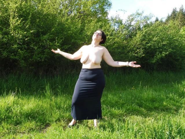 Topless EinfachDani-hot with big tits, wearing a dark skirt, standing in a grassy field, arms out, head tilted back, sunlight on her face, green trees in background.