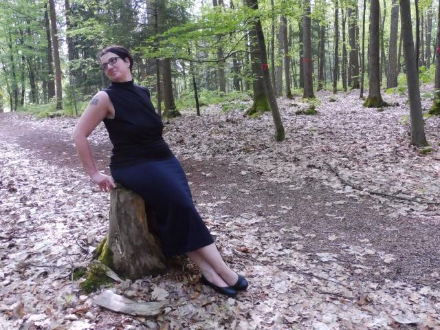 EinfachDani-hot, black sleeveless top, navy skirt, black flats, short dark hair, glasses, tattoo on right arm, leaning on tree stump, forest floor, leaves, moss, standing, legs crossed.
