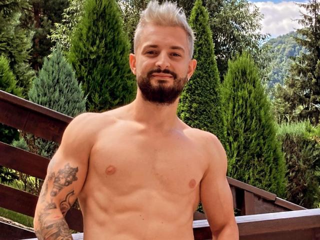 Meddelin, shirtless with blond, spiked hair, beard, tattoos on left arm, standing on wooden bridge, green trees, mountains in background, smiling, showing a toned, muscular, white chest.
