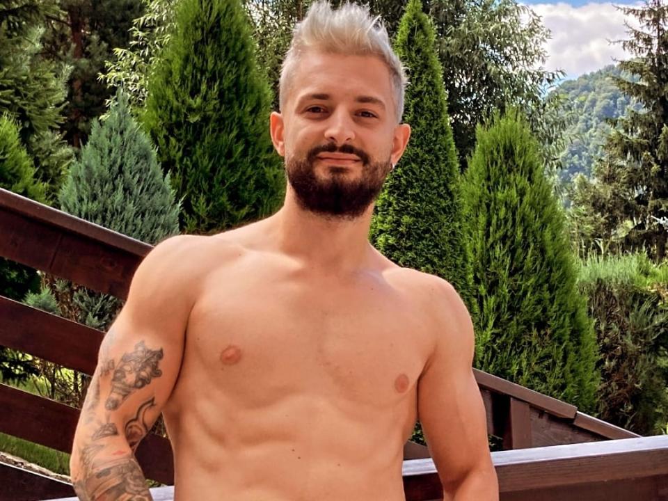 Meddelin, shirtless with blond, spiked hair, beard, tattoos on left arm, standing on wooden bridge, green trees, mountains in background, smiling, showing a toned, muscular, white chest.