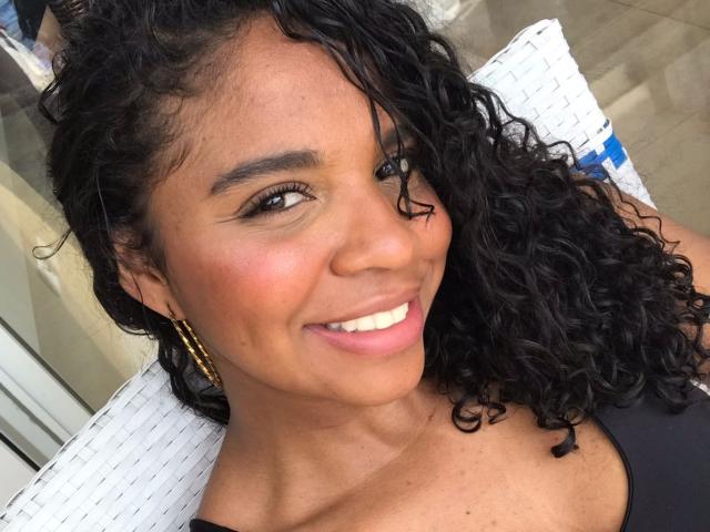 LisaMendez has curly black hair, brown skin, and a smiling face with white teeth, wearing gold earrings and a black top, looking to the side.