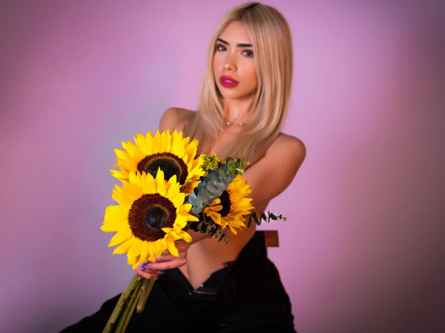 ChristieGarci with blonde hair, pink lipstick, holding sunflowers, wearing black top, exposing her tits, sitting, looking at camera, purple-pink background, soft lighting, sexy, pretty.