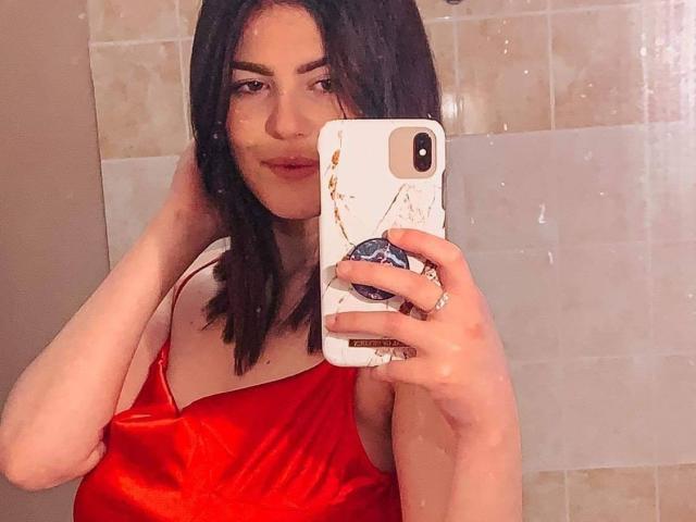 Trixie, con i capelli neri lunghi fino alle spalle, indossa un abito di seta rossa e sta scattando un selfie allo specchio con il cellulare. La sua mano tocca la testa, mostrando un sorriso appena accennato..