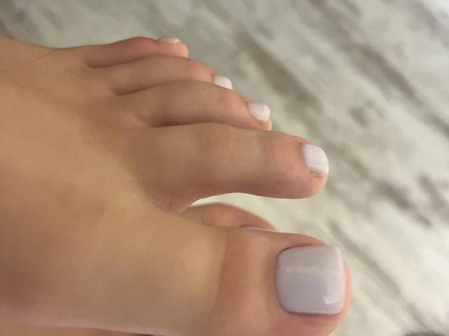 Destinis' foot with shiny white-painted toenails, showing her big toe and slightly curled toes, has smooth, light brown skin. The focus is on her painted toes in a tight shot.