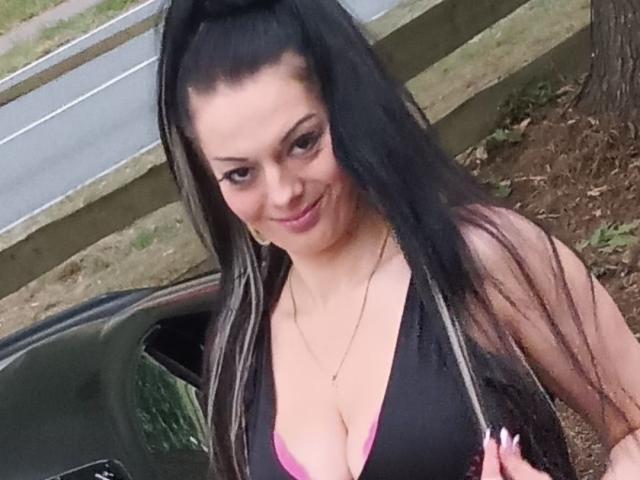 LuxusLady with long black hair in a high ponytail, smiling, wearing a black top with pink bra visible, showing her large tits, standing near a green car.