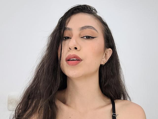 EmmaWells has long, dark, wet-looking hair, black eyeliner wings, red lipstick, and a small black earring in her right ear. She's wearing a black strap.