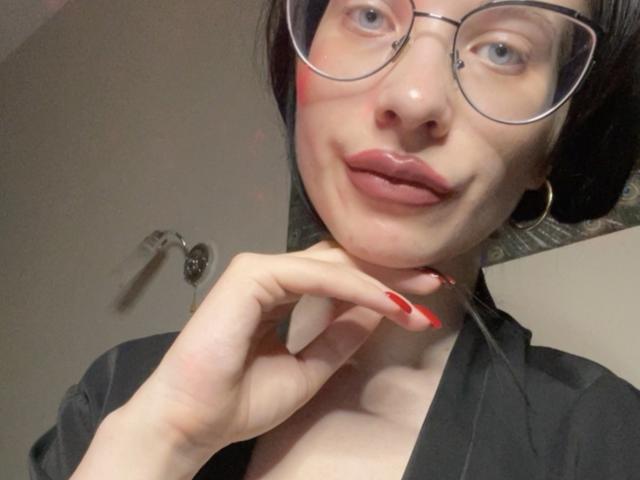 BelllaSd with glasses, red lipstick, and red nails, touching her chin, wearing a black shirt, has a cute smile, and is looking at the camera.