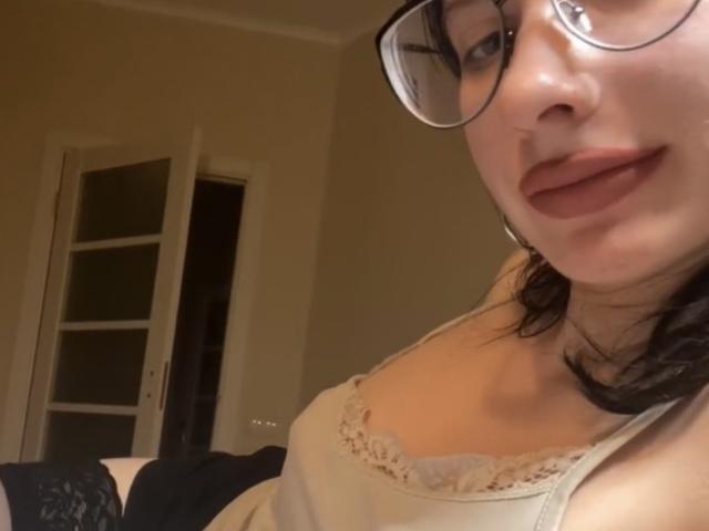 BelllaSd with glasses, pale skin, and dark hair, wearing a beige lace-trimmed top, showing her tits in a selfie, slightly smiling, in a dimly lit room.