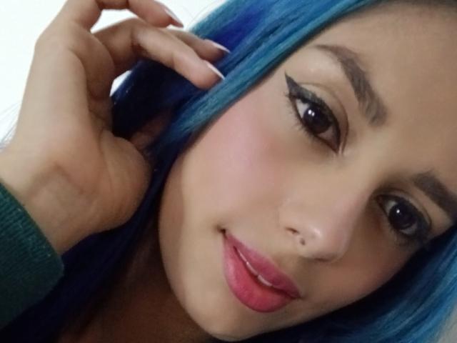AnaisHilton has blue hair, black eyeliner, pink lipstick, a nose piercing, and is looking at the camera with a slight smile, touching her ear with her hand.