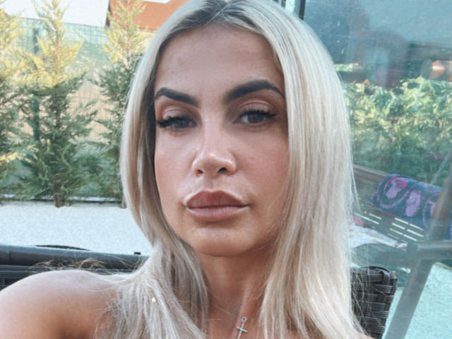 Lellie has blonde hair, thick eyebrows, and full lips with a cross necklace. Her blue eyes are slightly open, and she's giving a neutral expression. Boobs not visible.