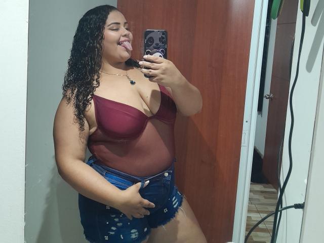 MelannieCarter with curly black hair, tongue out, wearing a sheer red bra and blue ripped denim shorts, holding a phone in a mirror selfie, showing her big tits.