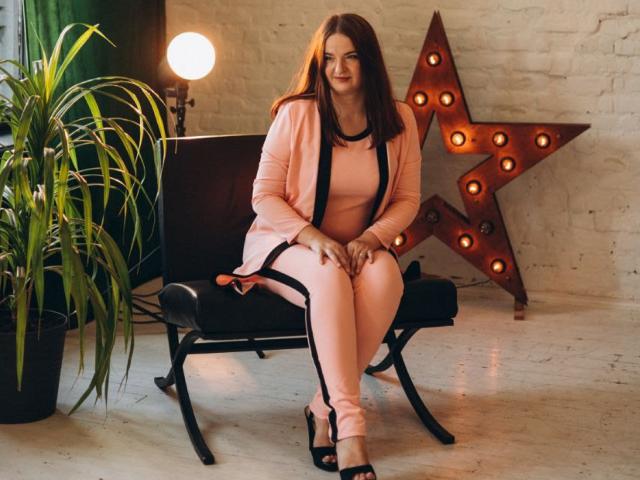 FreyaDak with brown hair, pink outfit, black strappy heels, sitting on black bench, hands on knees, white brick wall, large star-shaped light, green plant, and soft light.