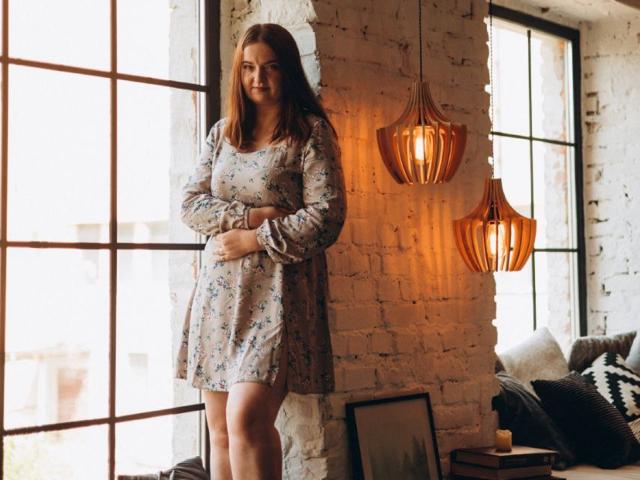 FreyaDak stands in a sunlit room, wearing a floral dress, arms crossed, brown hair, smiling, next to a white brick wall with hanging orange lanterns.