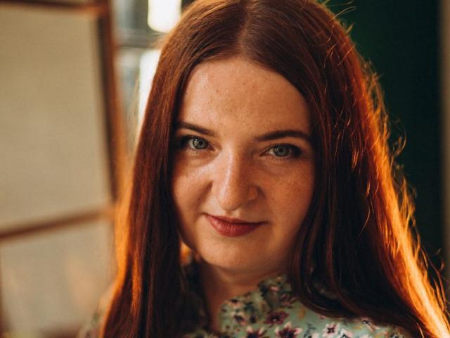 FreyaDak has long red hair, blue eyes, and a subtle smile with freckles on her face. She's wearing a floral shirt, and sunlight highlights her hair.