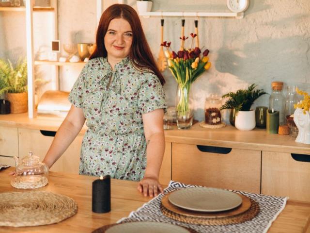 FreyaDak with red hair, wearing a floral dress, stands in a kitchen, smiling, holding a glass jar, wooden countertop, wicker placemat, plates, and a vase of yellow and purple tulips.
