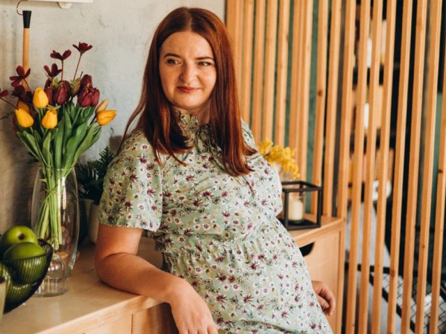 FreyaDak, com cabelo ruivo, vestido floral e um sorriso adorável, apoiada em um balcão de madeira com tulipas amarelas e roxas, uma maçã verde e ripas de madeira ao fundo..