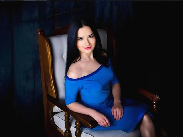 JudyLaVida with pale skin, long black hair, blue off-shoulder dress, sitting in a wooden chair, dark background, slight smile, red lipstick, visible thigh, relaxed posture.