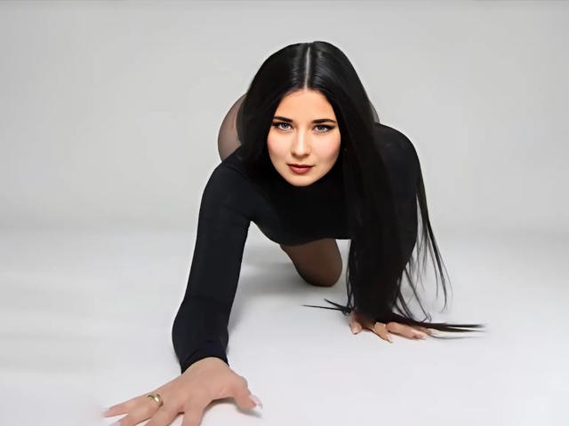 JudyLaVida, with long black hair, on all fours, wearing a black long-sleeve top, reaching forward with a gold ring on her finger, looking at the camera.