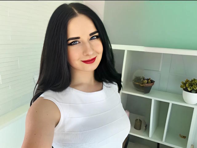 JudyLaVida with black hair, blue eyes, red lipstick, white sleeveless top, showing her tits, smiling, standing near a white shelf with plants.