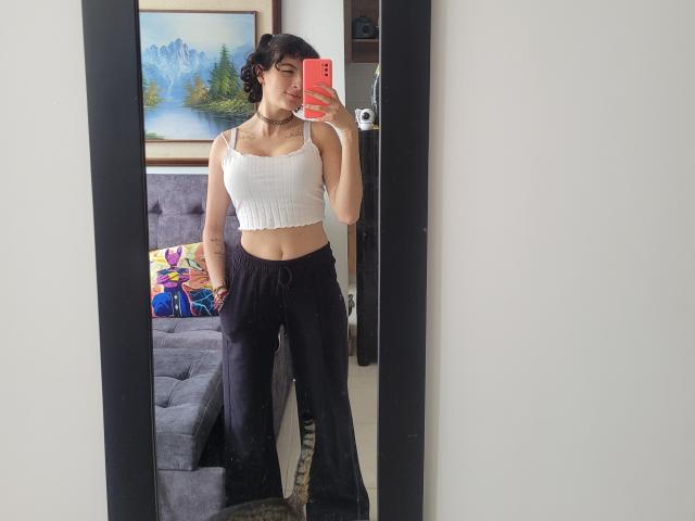 GabrielaMoore with curly dark hair, wearing a white ribbed crop top and black flared pants, taking a mirror selfie with an orange phone, showing small tits and a choker.