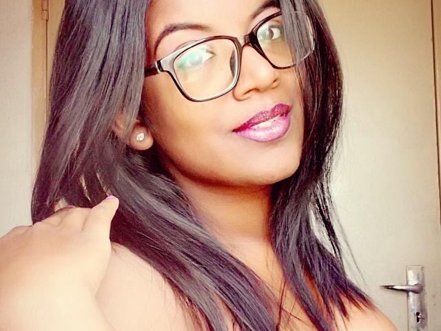 IndianBeaut with dark straight hair, glasses, shiny purple lipstick, and earrings, touching her hair, smiling. Her hand is near her head. She's wearing a strapless top.